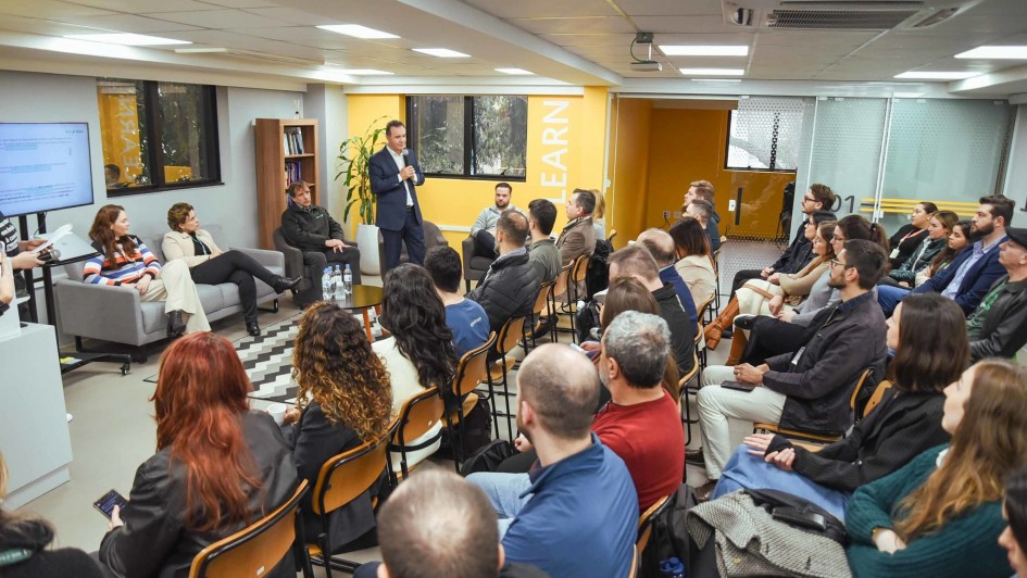 Esta é uma fotografia de um evento de treinamento ou apresentação em um ambiente de escritório ou sala de aula.
Em primeiro plano, a plateia é vista de costas, ocupando a maior parte da imagem. Homens e mulheres de diversas idades estão sentados em cadeiras de madeira, voltados para a frente da sala.
No centro da imagem, em pé, um homem de barba, usando uma jaqueta escura sobre uma camisa preta, está falando para o grupo. Ele parece estar segurando um pequeno controle remoto ou um microfone.
À esquerda, no fundo, uma mulher com cabelo escuro, vestindo uma camiseta preta, está de pé ao lado de uma tela de televisão grande que exibe um slide de apresentação com dados em colunas.
As paredes da sala são de cor cinza e amarela, com a palavra "LEARN" (aprender, em inglês) escrita em letras grandes e verticais em um pilar à direita. Há grandes janelas ao fundo da sala. O teto é branco e possui luminárias retangulares. A iluminação é clara e a imagem é nítida.