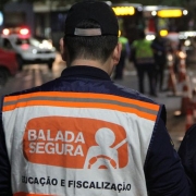 A foto mostra dois agentes de trânsito de costas em uma rua durante a noite. O agente à esquerda, um homem de boné preto, veste um colete branco com faixas laranjas refletivas, onde se lê "BALADA SEGURA" e "EDUCAÇÃO E FISCALIZAÇÃO" em letras pretas. A agente à direita, uma mulher de cabelos loiros, veste um casaco escuro com um distintivo "OPERAÇÃO BALADA SEGURA" nas costas. Ao fundo, carros com luzes de farol acesas e luzes vermelhas de emergência são visíveis, indicando que se trata de uma operação de trânsito em uma rua movimentada. A iluminação é proveniente dos veículos e das luzes da rua.