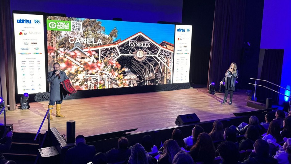 Foto de um evento realizado em um auditório com palco, onde duas pessoas estão se apresentando para uma plateia. À esquerda, um homem veste traje típico gaúcho, incluindo bota amarela, bombacha, casaco escuro e segura uma manta vermelha e preta. À direita, uma mulher com cabelos longos loiros fala ao microfone, vestindo roupas escuras e botas. Ao fundo, há um grande telão digital com a imagem do pórtico da cidade de Canela (RS), acompanhado dos dizeres “CANELA” e “Viva o Inverno Gaúcho”. Nas laterais do telão, banners com logomarcas de patrocinadores ou apoiadores do evento. A plateia está atenta, com pessoas vestindo casacos, indicando clima frio. O ambiente tem iluminação azul e roxa suave.