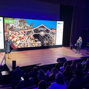 Foto de um evento realizado em um auditório com palco, onde duas pessoas estão se apresentando para uma plateia. À esquerda, um homem veste traje típico gaúcho, incluindo bota amarela, bombacha, casaco escuro e segura uma manta vermelha e preta. À direita, uma mulher com cabelos longos loiros fala ao microfone, vestindo roupas escuras e botas. Ao fundo, há um grande telão digital com a imagem do pórtico da cidade de Canela (RS), acompanhado dos dizeres “CANELA” e “Viva o Inverno Gaúcho”. Nas laterais do telão, banners com logomarcas de patrocinadores ou apoiadores do evento. A plateia está atenta, com pessoas vestindo casacos, indicando clima frio. O ambiente tem iluminação azul e roxa suave.