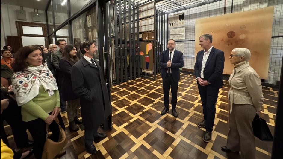 A foto mostra um ambiente interno que parece ser uma área de armazenamento de obras de arte do museu. Ao fundo, o Secretário da Cultura, Eduardo Loureiro, de terno e gravata, está em pé e sorri levemente. Ele está à frente de um homem que é o diretor-curador do MARGS, Francisco Dalcol, que usa barba, óculos e uma blusa escura, e também está sorrindo. Ambos estão à frente de uma obra de arte grande, parcialmente coberta por um tecido branco. A sala tem paredes brancas e está cheia de grandes telas e outras peças de arte dispostas em cavaletes e prateleiras.