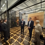 A foto mostra um ambiente interno que parece ser uma área de armazenamento de obras de arte do museu. Ao fundo, o Secretário da Cultura, Eduardo Loureiro, de terno e gravata, está em pé e sorri levemente. Ele está à frente de um homem que é o diretor-curador do MARGS, Francisco Dalcol, que usa barba, óculos e uma blusa escura, e também está sorrindo. Ambos estão à frente de uma obra de arte grande, parcialmente coberta por um tecido branco. A sala tem paredes brancas e está cheia de grandes telas e outras peças de arte dispostas em cavaletes e prateleiras.