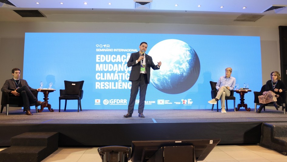 Esta é uma fotografia de um painel de discussão em um seminário.
No centro da imagem, em um palco elevado, um homem com barba e vestindo um terno escuro está em pé, no meio de outras três pessoas sentadas. Ele gesticula com as mãos enquanto fala para uma plateia que não está visível.
Atrás do palco, um grande telão azul exibe o título "SEMINÁRIO INTERNACIONAL" e as palavras "EDUCAÇÃO, MUDANÇAS CLIMÁTICAS e RESILIÊNCIA", junto com logotipos de organizações como "GFDRR" e "WORLD BANK".
No palco, à esquerda do orador, um homem de terno escuro está sentado em uma poltrona preta, com as pernas cruzadas. À direita do orador, um homem de cabelo claro e camisa social está sentado, e ao seu lado, uma mulher de cabelo escuro, vestindo uma blusa preta, também está sentada.
O chão do palco é preto e há mesas de apoio com garrafas de água. A iluminação é focada no palco, destacando os participantes.