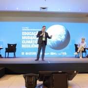 Esta é uma fotografia de um painel de discussão em um seminário.

No centro da imagem, em um palco elevado, um homem com barba e vestindo um terno escuro está em pé, no meio de outras três pessoas sentadas. Ele gesticula com as mãos enquanto fala para uma plateia que não está visível.

Atrás do palco, um grande telão azul exibe o título "SEMINÁRIO INTERNACIONAL" e as palavras "EDUCAÇÃO, MUDANÇAS CLIMÁTICAS e RESILIÊNCIA", junto com logotipos de organizações como "GFDRR" e "WORLD BANK".

No palco, à esquerda do orador, um homem de terno escuro está sentado em uma poltrona preta, com as pernas cruzadas. À direita do orador, um homem de cabelo claro e camisa social está sentado, e ao seu lado, uma mulher de cabelo escuro, vestindo uma blusa preta, também está sentada.

O chão do palco é preto e há mesas de apoio com garrafas de água. A iluminação é focada no palco, destacando os participantes.
