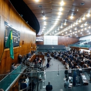 A foto mostra o plenário da Assembleia Legislativa do Rio Grande do Sul, um grande salão com paredes de madeira escura e um teto alto com iluminação moderna. No primeiro plano, uma escadaria leva a um andar superior, com um corrimão de vidro. No centro da sala, há um auditório com várias fileiras de cadeiras estofadas escuras, onde a plateia está sentada. Em uma área mais elevada, à esquerda, uma longa mesa retangular com cadeiras acomoda um grupo de pessoas, que parecem ser membros da mesa diretora ou de uma comissão. Ao fundo, uma grande tela de LED exibe informações. O ambiente está bem iluminado, e muitas pessoas, em sua maioria em trajes formais, estão presentes na sessão.