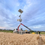 Foto ao ar livre, em um dia claro. Há uma estação meteorológica em cima de uma caminhão no meio de um campo. Por perto, há duas pessoas olhando para o equipamento. 
