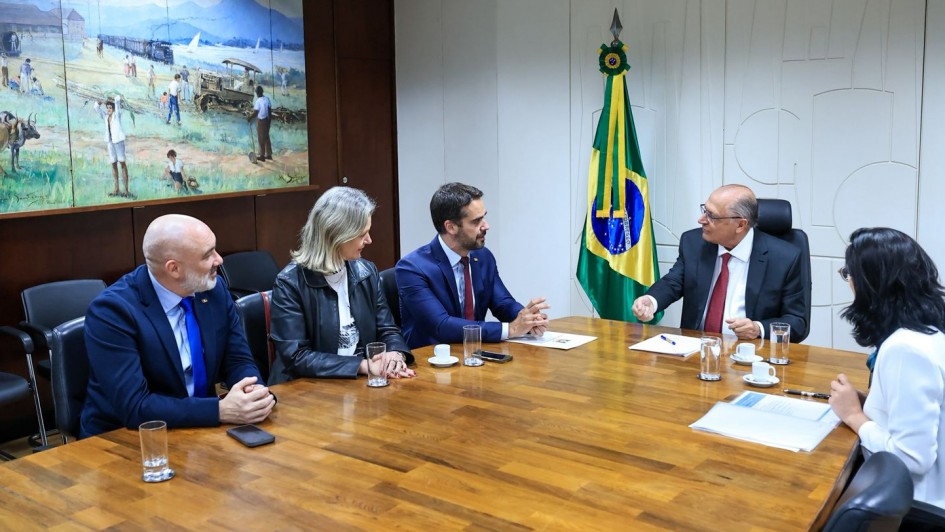 Foto de uma sala de reunião formal, com cinco pessoas sentadas ao redor de uma mesa de maneira. Leite aparece ao centro, olhando para Alkmin, que fala.