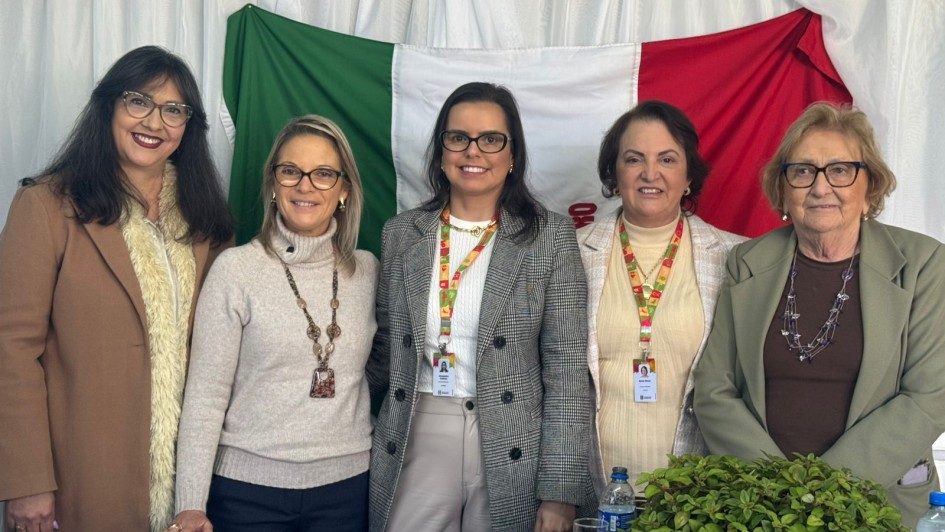 Esta é uma fotografia de um grupo de cinco mulheres, de pé atrás de uma mesa, posando para a câmera.
Ao fundo, há uma grande bandeira estendida, com três faixas verticais nas cores verde, branca e vermelha, e o que parece ser um logotipo no centro. Na frente do grupo, há uma mesa coberta com uma toalha branca. Sobre a mesa, em primeiro plano, há um arranjo de folhas verdes e garrafas de água.
Da esquerda para a direita, as mulheres estão vestidas da seguinte forma:
Uma mulher com cabelos escuros e óculos, vestindo um casaco marrom com gola de pele clara.
Uma mulher loira de óculos, vestindo um suéter bege de gola alta.
Uma mulher com cabelos escuros e óculos, vestindo um blazer xadrez cinza sobre uma blusa branca.
Uma mulher com cabelos escuros e um colar de pérolas, vestindo um suéter de gola alta creme e um cordão de crachá no pescoço.
Uma mulher mais velha com cabelo curto e óculos, vestindo um blazer verde-claro.
Todas as mulheres estão sorrindo para a foto. A iluminação é clara e uniforme.