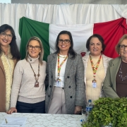 Esta é uma fotografia de um grupo de cinco mulheres, de pé atrás de uma mesa, posando para a câmera.

Ao fundo, há uma grande bandeira estendida, com três faixas verticais nas cores verde, branca e vermelha, e o que parece ser um logotipo no centro. Na frente do grupo, há uma mesa coberta com uma toalha branca. Sobre a mesa, em primeiro plano, há um arranjo de folhas verdes e garrafas de água.

Da esquerda para a direita, as mulheres estão vestidas da seguinte forma:

Uma mulher com cabelos escuros e óculos, vestindo um casaco marrom com gola de pele clara.

Uma mulher loira de óculos, vestindo um suéter bege de gola alta.

Uma mulher com cabelos escuros e óculos, vestindo um blazer xadrez cinza sobre uma blusa branca.

Uma mulher com cabelos escuros e um colar de pérolas, vestindo um suéter de gola alta creme e um cordão de crachá no pescoço.

Uma mulher mais velha com cabelo curto e óculos, vestindo um blazer verde-claro.

Todas as mulheres estão sorrindo para a foto. A iluminação é clara e uniforme.