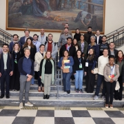 Esta é uma fotografia de um grande grupo de pessoas, aproximadamente 30, posando para uma foto em uma escadaria de um prédio.

O grupo é formado por homens e mulheres de diversas idades, a maioria vestida com roupas sociais ou casuais. Eles estão dispostos em vários degraus de uma larga escadaria, olhando e sorrindo para a câmera. Alguns estão segurando cadernos, pastas ou outros materiais, e muitos usam crachás de identificação.

Ao fundo, há uma grande pintura emoldurada pendurada na parede, representando uma cena histórica ou rural com várias figuras humanas. O chão no pé da escada é um piso xadrez preto e branco. O ambiente é formal e a iluminação é clara.