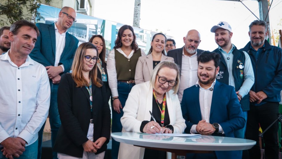 A foto mostra o momento de assinatura do documento. A Secretária Arita Bergmann, com óculos e um casaco branco, está em pé, sorrindo, enquanto assina um papel em uma mesa redonda e branca. Ao seu lado, um homem de terno azul-marinho e um homem de camisa branca e calça jeans a observam. Outras pessoas, incluindo um homem com uma camisa polo preta e uma mulher com um blazer branco, estão em volta, sorrindo e assistindo ao ato. O evento acontece em uma área externa, sob uma cobertura, com a presença de outras pessoas ao fundo.