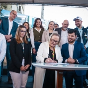 A foto mostra o momento de assinatura do documento. A Secretária Arita Bergmann, com óculos e um casaco branco, está em pé, sorrindo, enquanto assina um papel em uma mesa redonda e branca. Ao seu lado, um homem de terno azul-marinho e um homem de camisa branca e calça jeans a observam. Outras pessoas, incluindo um homem com uma camisa polo preta e uma mulher com um blazer branco, estão em volta, sorrindo e assistindo ao ato. O evento acontece em uma área externa, sob uma cobertura, com a presença de outras pessoas ao fundo.