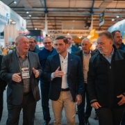 Um grupo de homens caminha por um corredor coberto de uma feira ou evento. No centro da imagem, três deles estão em destaque, conversando entre si. O homem do meio veste blazer azul-marinho, camisa branca e calça clara; ele parece liderar a comitiva. À esquerda, outro homem calvo com terno cinza e crachá conversa com ele, gesticulando com as mãos. À direita, um terceiro homem com barba grisalha veste jaqueta escura e observa a conversa. Ao fundo, outras pessoas acompanham o grupo e há estandes iluminados, com logotipos visíveis e iluminação artificial no teto, sugerindo um ambiente profissional e organizado, como uma feira de negócios ou exposição industrial.