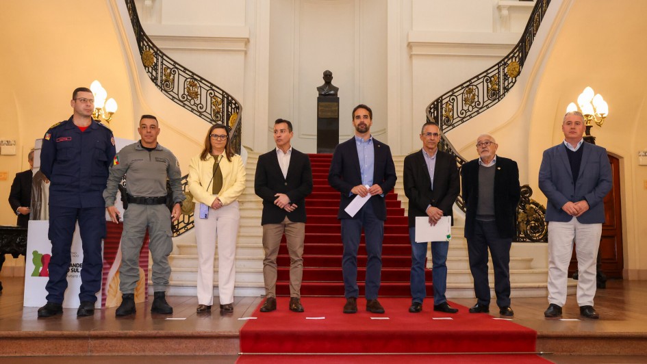 Grupo de oito pessoas está posicionado lado a lado sobre um tapete vermelho, no centro de uma escadaria interna com corrimões ornamentados, em um ambiente de arquitetura clássica. Entre os presentes, há homens e uma mulher, alguns em trajes formais (terno e gravata), outros em uniformes militares, como policiais e bombeiros. Ao fundo, observa-se um busto em pedestal e paredes com detalhes decorativos. O local é bem iluminado, com lustres antigos nas laterais. Trata-se de um evento oficial, possivelmente relacionado ao governo ou à segurança pública.