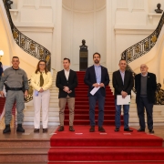 Grupo de oito pessoas está posicionado lado a lado sobre um tapete vermelho, no centro de uma escadaria interna com corrimões ornamentados, em um ambiente de arquitetura clássica. Entre os presentes, há homens e uma mulher, alguns em trajes formais (terno e gravata), outros em uniformes militares, como policiais e bombeiros. Ao fundo, observa-se um busto em pedestal e paredes com detalhes decorativos. O local é bem iluminado, com lustres antigos nas laterais. Trata-se de um evento oficial, possivelmente relacionado ao governo ou à segurança pública.
