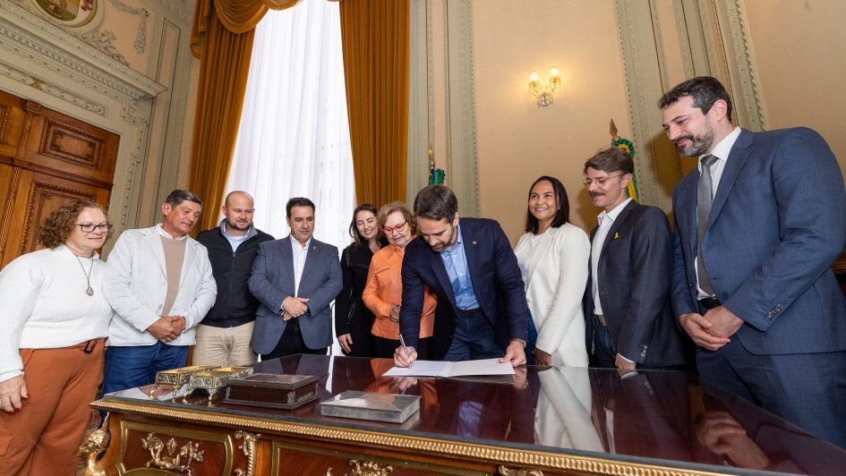 A imagem mostra o governador do Rio Grande do Sul, Eduardo Leite, sancionando projetos de lei. Ele está no centro da foto, inclinado sobre uma grande mesa de madeira escura com detalhes dourados, assinando um documento. Ele usa um terno azul-marinho.

Ao redor da mesa, estão várias pessoas. À sua direita (nossa esquerda), há um grupo de cinco pessoas: duas mulheres, um homem mais velho com chapéu e dois outros homens. A mulher mais próxima ao governador tem cabelos loiros e usa uma blusa laranja. A mulher na extremidade esquerda, com óculos, usa uma blusa branca de manga comprida e calças de cor de tijolo.

À esquerda do governador (nossa direita), estão três pessoas: duas mulheres e um homem. A mulher mais próxima ao governador, com cabelos escuros e lisos, usa uma blusa branca. Ao lado dela, está um homem com bigode e cabelo mais longo, usando um terno. Na extremidade direita, há um homem alto, com barba, usando um terno azul, olhando para o governador com um sorriso.

O ambiente parece ser um gabinete ou uma sala de reuniões oficial, com cortinas pesadas e uma estante de madeira ao fundo. A iluminação é clara, e a atmosfera parece ser de um evento formal e positivo.