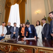 A imagem mostra o governador do Rio Grande do Sul, Eduardo Leite, sancionando projetos de lei. Ele está no centro da foto, inclinado sobre uma grande mesa de madeira escura com detalhes dourados, assinando um documento. Ele usa um terno azul-marinho.

Ao redor da mesa, estão várias pessoas. À sua direita (nossa esquerda), há um grupo de cinco pessoas: duas mulheres, um homem mais velho com chapéu e dois outros homens. A mulher mais próxima ao governador tem cabelos loiros e usa uma blusa laranja. A mulher na extremidade esquerda, com óculos, usa uma blusa branca de manga comprida e calças de cor de tijolo.

À esquerda do governador (nossa direita), estão três pessoas: duas mulheres e um homem. A mulher mais próxima ao governador, com cabelos escuros e lisos, usa uma blusa branca. Ao lado dela, está um homem com bigode e cabelo mais longo, usando um terno. Na extremidade direita, há um homem alto, com barba, usando um terno azul, olhando para o governador com um sorriso.

O ambiente parece ser um gabinete ou uma sala de reuniões oficial, com cortinas pesadas e uma estante de madeira ao fundo. A iluminação é clara, e a atmosfera parece ser de um evento formal e positivo.