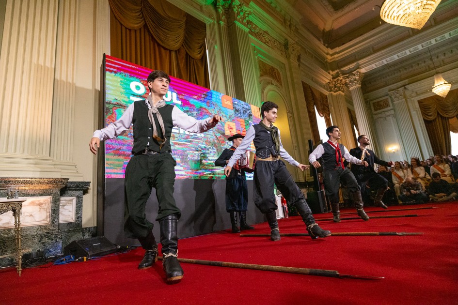 Foto, meio de lado, de uma apresentação de dança típica no interior do Palácio Piratini. Quatro homens em trajes típicos gaúchos aparecem sapateando. Um músico toca sanfona ao fundo.