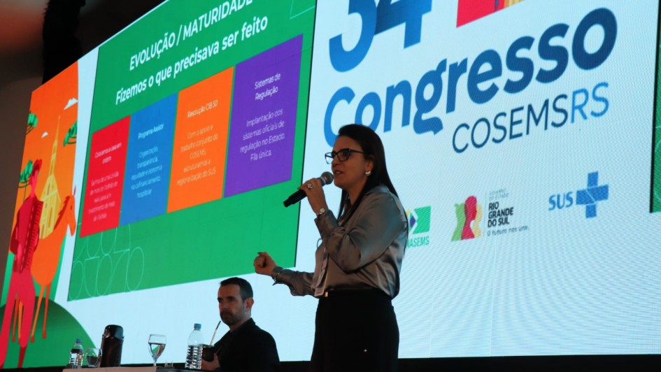 Foto de Lisiane ao centro, meio de lado, falando com o microfone na mão. Ao fundo, há um painel com a logo do evento.