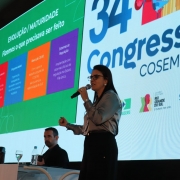 Foto de Lisiane ao centro, meio de lado, falando com o microfone na mão. Ao fundo, há um painel com a logo do evento.