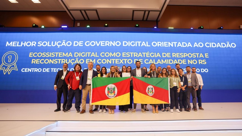 A imagem mostra um grupo de pessoas reunidas em um palco, segurando duas bandeiras do estado do Rio Grande do Sul, posando para a foto. Ao fundo, há um grande painel azul com o texto: “Melhor solução de governo digital orientada ao cidadão. Ecossistema digital como estratégia de resposta e reconstrução em contexto de calamidade no RS."