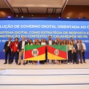 A imagem mostra um grupo de pessoas reunidas em um palco, segurando duas bandeiras do estado do Rio Grande do Sul, posando para a foto. Ao fundo, há um grande painel azul com o texto: “Melhor solução de governo digital orientada ao cidadão. Ecossistema digital como estratégia de resposta e reconstrução em contexto de calamidade no RS."