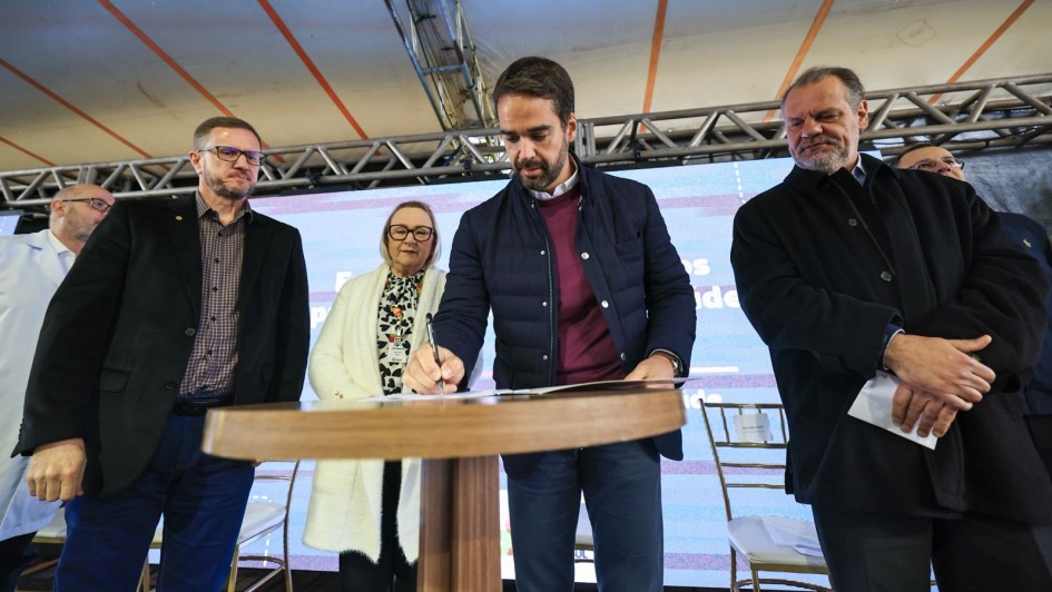 A foto mostra o governador Eduardo Leite assinando um documento com uma caneta. Ele está em pé atrás de mesa redonda de madeira claro com outras pessoas ao redor, em um ambiente externo e coberto.