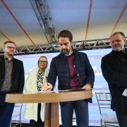 A foto mostra o governador Eduardo Leite assinando um documento com uma caneta. Ele está em pé atrás de mesa redonda de madeira claro com outras pessoas ao redor, em um ambiente externo e coberto.