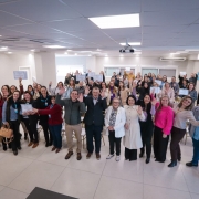 A imagem mostra um grupo grande de pessoas reunidas em um ambiente interno claro e moderno, possivelmente uma sala de eventos ou auditório. Elas estão posicionadas de frente para a câmera, muitas sorrindo e acenando com uma das mãos levantadas. Algumas seguram certificados, indicando que o momento é de celebração ou formatura. As cadeiras ao fundo estão parcialmente ocupadas, e há um projetor no teto. O grupo é diverso, composto majoritariamente por mulheres, e todos parecem estar vestidos de forma casual ou social.