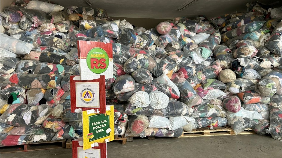 A foto mostra um grande amontoado de roupas e agasalhos ensacados, que preenche a maior parte da imagem. As doações estão organizadas em sacos plásticos transparentes e são empilhadas sobre pallets. Em primeiro plano, há um banner de papelão em formato de totem, com informações sobre a "Campanha do Agasalho" e a mensagem "Faça sua doação aqui".