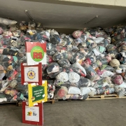 A foto mostra um grande amontoado de roupas e agasalhos ensacados, que preenche a maior parte da imagem. As doações estão organizadas em sacos plásticos transparentes e são empilhadas sobre pallets. Em primeiro plano, há um banner de papelão em formato de totem, com informações sobre a "Campanha do Agasalho" e a mensagem "Faça sua doação aqui".