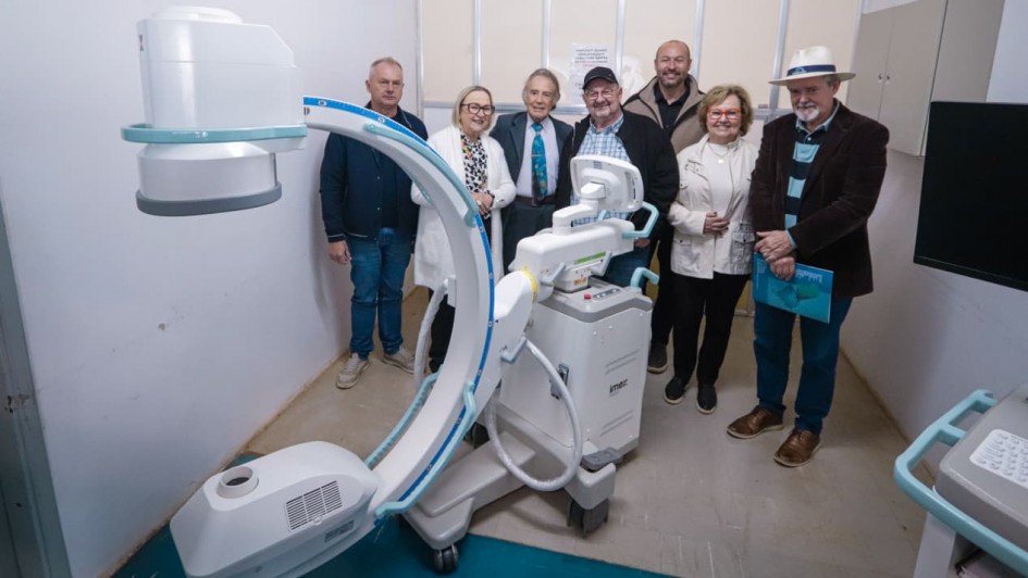 A foto mostra um grupo de sete pessoas, incluindo a secretária da Saúde, Arita Bergmann, em uma sala com paredes brancas. O grupo está posicionado ao redor de um grande equipamento médico de cor clara, com braços articulados e rodinhas, que está no centro da imagem. Arita Bergmann está no centro da foto, usando um casaco branco e sorrindo. Todos olham para a câmera.