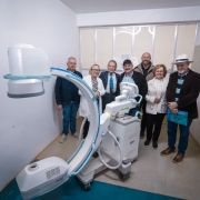 A foto mostra um grupo de sete pessoas, incluindo a secretária da Saúde, Arita Bergmann, em uma sala com paredes brancas. O grupo está posicionado ao redor de um grande equipamento médico de cor clara, com braços articulados e rodinhas, que está no centro da imagem. Arita Bergmann está no centro da foto, usando um casaco branco e sorrindo. Todos olham para a câmera.