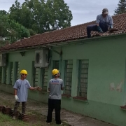 A foto mostra o telhado de um prédio escolar de um andar com telhas de cor terracota. Há três homens trabalhando na reforma do telhado. Um deles está no telhado, agachado e removendo as telhas, enquanto os outros dois estão no chão, de capacete amarelo, inspecionando o trabalho. As paredes do prédio são de cor verde-clara, e há aparelhos de ar-condicionado instalados na parte externa.
