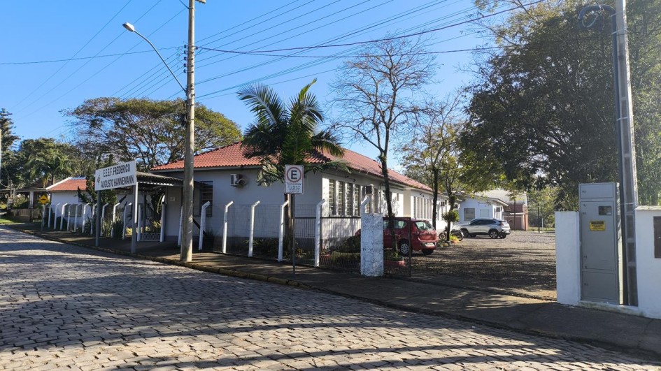 A foto é uma vista externa da Escola Frederico Augusto Hanemann, em um dia ensolarado. O prédio, de um andar, tem paredes brancas e telhado de telhas vermelhas. Há grandes janelas de vidro e uma placa com o nome da escola está visível na fachada. O prédio é cercado por um muro baixo e vegetação, e uma rua de paralelepípedos e um poste de luz podem ser vistos em primeiro plano.