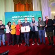 A imagem mostra um grupo de 11 pessoas, em pé, em um palco com um tapete vermelho. No centro, dois homens estão segurando um documento. Um deles está de terno, o outro está de blazer. Atrás deles, há um grande painel digital com o texto "ASSINATURA DA PORTARIA SES". As outras pessoas estão aplaudindo, algumas olhando para o documento, outras para o público. O cenário inclui cortinas de veludo e paredes decoradas.