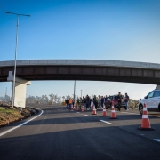 A imagem mostra um grupo de pessoas em pé em uma rodovia, próximo a um viaduto. O viaduto atravessa a rodovia na parte superior da imagem. Algumas pessoas parecem estar em uma cerimônia ou evento de inauguração. Há cones de trânsito laranja e carros estacionados, incluindo um SUV branco à direita e uma van branca ao fundo. O céu está limpo e azul.