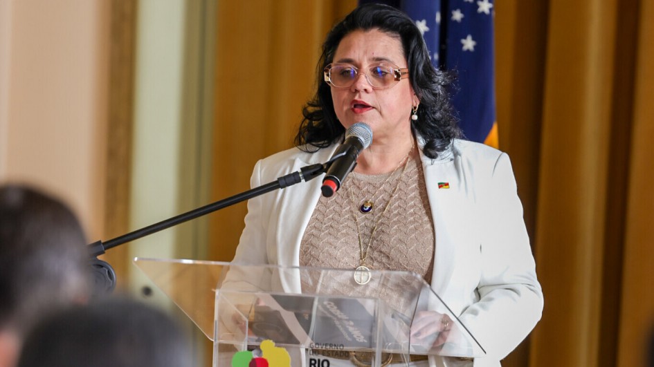A secretária do Meio Ambiente, Marjorie Kauffmann, com cabelo escuro e óculos de armação dourada, fala a um microfone em um púlpito de acrílico. Ela veste um blazer branco sobre uma blusa de cor clara e usa um colar com um pingente. No púlpito, a inscrição "CORSAN 2 ANOS" é visível. Ao fundo, uma bandeira do Rio Grande do Sul pode ser vista.