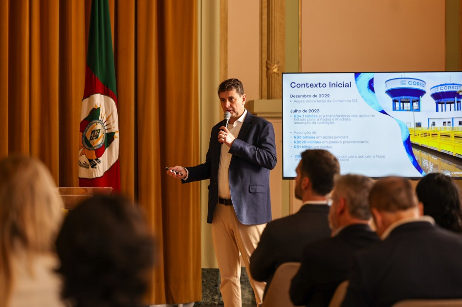 O vice-presidente de Operações do Grupo Aegea, Leandro Marin, de camisa social azul marinho e calças cáqui, está de pé em um palco, segurando um microfone e gesticulando com a mão esquerda em direção a um público. Ao fundo, uma tela de apresentação mostra o título "Contexto Inicial" e tópicos relacionados à Corsan, indicando que ele está fazendo uma apresentação. A bandeira do Rio Grande do Sul é visível à sua esquerda.