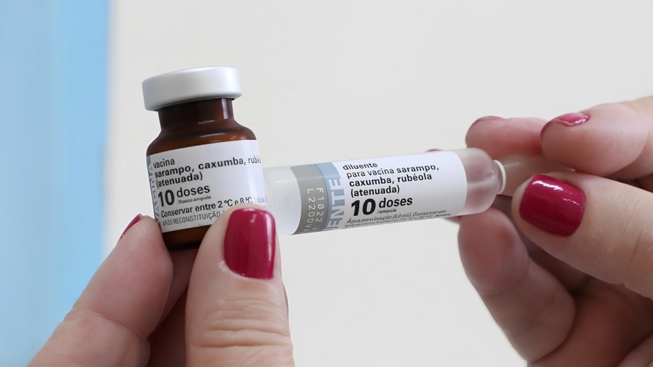 Imagem mostra um frasco de vidro marrom com tampa branca e um rótulo escrito “vacina sarampo, caxumba, rubéola (atenuada) – 10 doses” sendo segurado por uma mão. Na outra mão, há um frasco transparente com tampa branca, identificado como “diluente para vacina sarampo, caxumba, rubéola (atenuada) – 10 doses”. As unhas de ambas as mãos estão pintadas com esmalte vermelho.