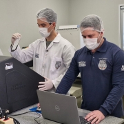 A imagem mostra dois homens, ambos de touca e máscara de proteção, em um laboratório. O homem da esquerda veste um jaleco branco e luvas, e segura um pequeno tubo de ensaio. O homem da direita veste uma camiseta azul escura com a inscrição "PERÍCIA CRIMINAL" e o símbolo da Polícia Federal. Ele está sentado em frente a um notebook Dell, com a mão direita sobre o mouse e a esquerda sobre o teclado, aparentemente interagindo com o computador.

Entre os dois, há um equipamento grande e preto com o logo "Applied Biosystems" e a sigla "FNSP" visível. O ambiente de fundo é um laboratório, com bancadas cinzas de granito, diversos outros equipamentos de laboratório e janelas com molduras brancas que separam o espaço de outro cômodo. O homem de jaleco tem cabelos escuros, e o de camiseta azul tem barba.