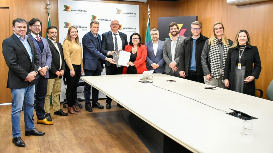 A foto mostra um grupo de onze pessoas posando para a câmera em uma sala de reuniões. No centro, um homem de terno e uma mulher de blazer vermelho estão segurando um documento. A mesa é grande e branca, e ao fundo há um painel com a inscrição "Rio Grande do Sul" e os logos do governo do Estado.