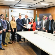 A foto mostra um grupo de onze pessoas posando para a câmera em uma sala de reuniões. No centro, um homem de terno e uma mulher de blazer vermelho estão segurando um documento. A mesa é grande e branca, e ao fundo há um painel com a inscrição "Rio Grande do Sul" e os logos do governo do Estado.