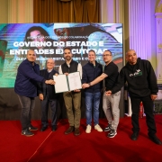 A foto mostra um grupo de seis homens, com o governador Eduardo Leite no centro, posando para a câmera em um palco. Eles estão segurando um grande certificado ou documento de cor verde. O ato ocorreu no Palácio Piratini.