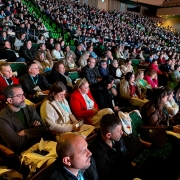 Auditório cheio, com muitas pessoas sentadas em cadeiras verdes, assistindo a um evento iluminado.