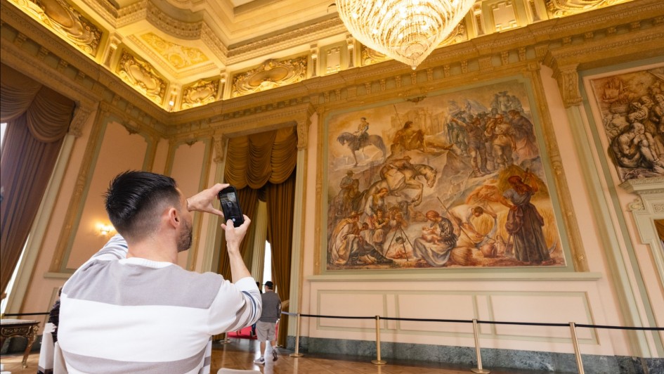 A foto mostra o interior de um salão do Palácio Piratini. Em primeiro plano, um homem de costas, vestindo uma camiseta listrada, está tirando uma foto com o celular de uma grande pintura mural do artista Aldo Locatelli. A obra de arte retrata uma batalha e ocupa uma parede inteira. O salão tem piso de madeira e paredes de cor clara, com detalhes em dourado e um lustre de cristal no teto.