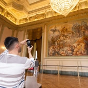 A foto mostra o interior de um salão do Palácio Piratini. Em primeiro plano, um homem de costas, vestindo uma camiseta listrada, está tirando uma foto com o celular de uma grande pintura mural do artista Aldo Locatelli. A obra de arte retrata uma batalha e ocupa uma parede inteira. O salão tem piso de madeira e paredes de cor clara, com detalhes em dourado e um lustre de cristal no teto.