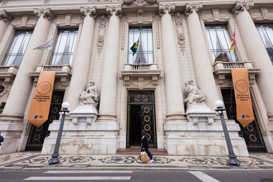 A foto mostra a fachada do Palácio Piratini. O prédio é de estilo neoclássico, com grandes colunas e janelas altas. Duas estátuas de figuras femininas e duas de figuras masculinas estão posicionadas em frente às colunas. Em cada lado da entrada principal, há banners alaranjados com o texto "Dia Estadual do Patrimônio Cultural". Em primeiro plano, um pedestre está caminhando na calçada em frente ao prédio.