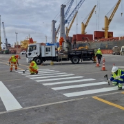 Descrição para acessibilidade: Trabalhadores com coletes refletivos e capacetes realizam a pintura de uma faixa de pedestres em área portuária. Ao fundo, há um caminhão de apoio, cones de sinalização, guindastes e grandes navios de carga atracados, com contêineres e equipamentos de operação portuária visíveis. O céu está nublado.