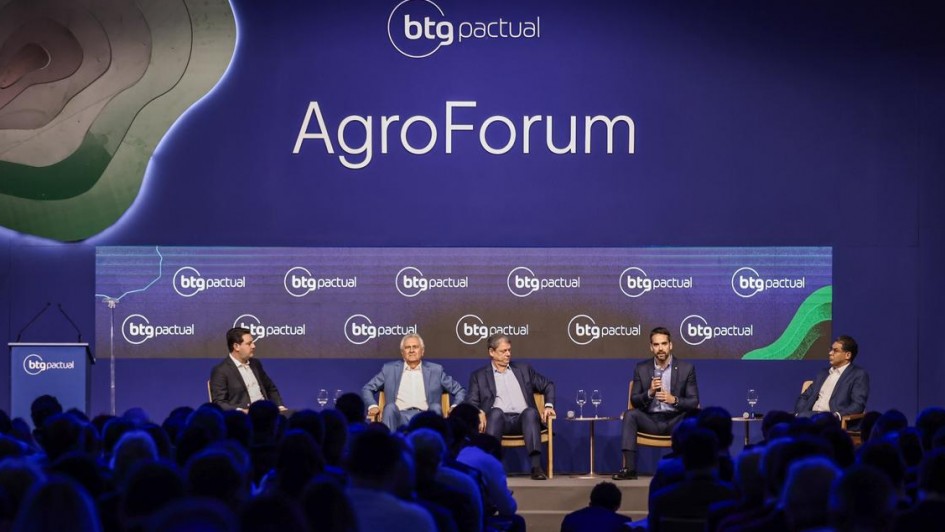 A foto mostra cinco homens sentados em um palco, participando de um painel. O governador do RS, Eduardo Leite, fala ao microfone. A plateia acompanha a atividade. Ao fundo há inscrições repetidas de "btg pactual" e, em tamanho maior, a palavra "AgroForum". 